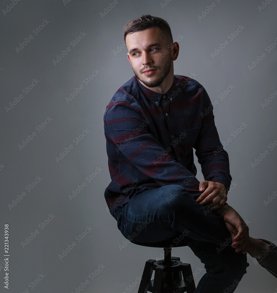 Handsome young thoughtful man in checkered shirt sitting on the chair and looking at camera isolated on a gray background