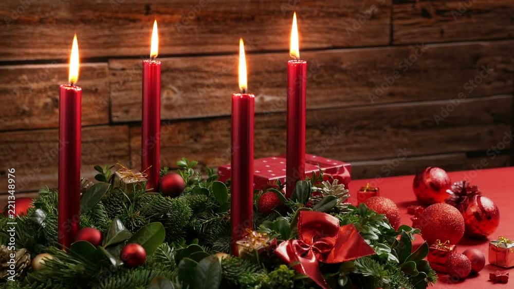 Four burning red candles on a traditional advent wreath of green fir twigs and mistletoes with festive decoration in front of a rustic wooden wall, close-up real time shot with copy space, nobody