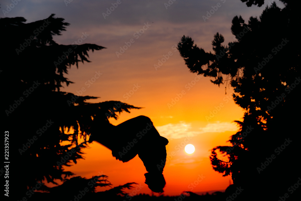 Silhouette of young parkour man while jumping and flipping upside down in park on a sunrise. 