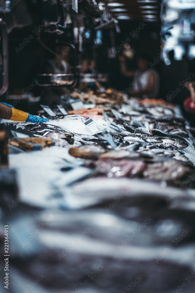 The counter on the mart with raw fish surrounded by grated ice, shallow depth of field; a street market with fish-shop, strong bokeh, people in a defocused background, Barcelona, Spain