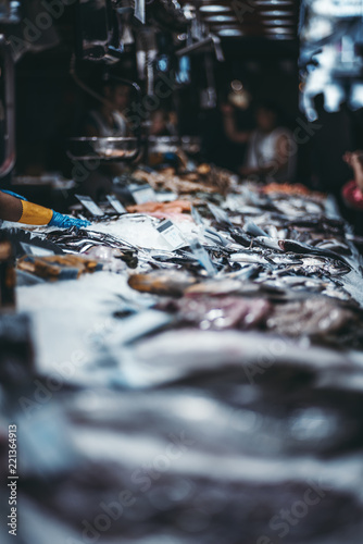 The counter on the mart with raw fish surrounded by grated ice, shallow depth of field; a street market with fish-shop, strong bokeh, people in a defocused background, Barcelona, Spain