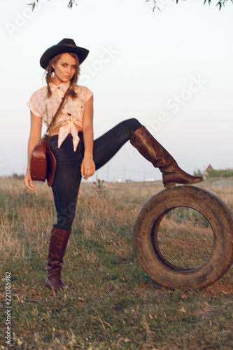 Girl cowgirl in jeans boots shirt and with a bag in a box with a wheel by a tree