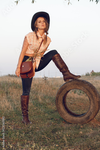 Girl cowgirl in jeans boots shirt and with a bag in a box with a wheel by a tree