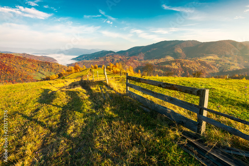 beautiful countryside at foggy autumn sunrise. wooden fence across the rural alpine meadow red foliage on forested hills. cloud inversion in distant valley. beautiful season colors