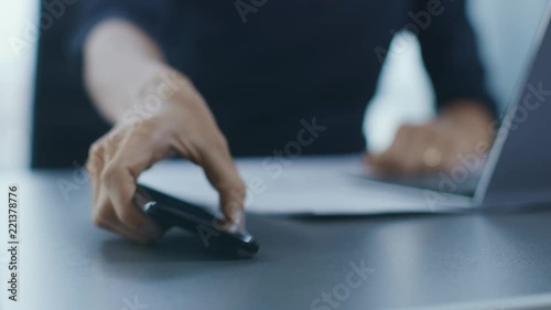 Out of Focus Shot of the Businesswoman Working at Her Office Desk Reaching Out for Her Smartphone and Starts Using It. Woman Picks up Mobile Phone from Her Desk. Focus on a Phone.