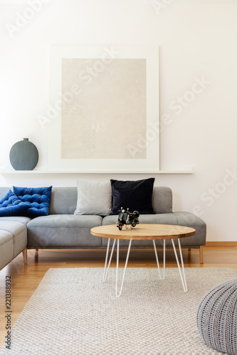 Gray poster on a shelf above a gray corner sofa with pillows in a simple living room interior with white walls