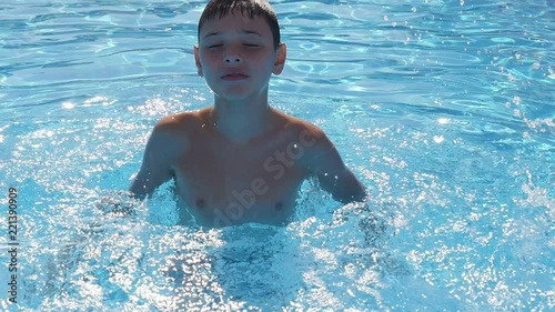 A cheerful view of a small boy enjoying life in a swimming pool with sky blue waters in summer in slo-mo. He makes a lot of splashes and feels fine  photo