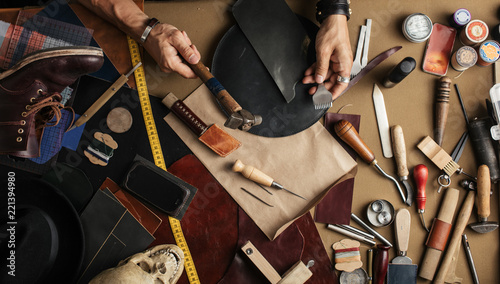 High angle view of craftsman at work over background with set of cobbler tools and colored leather in rolls. Genuine leather handbag master at work in local workshop. Handmade concept.