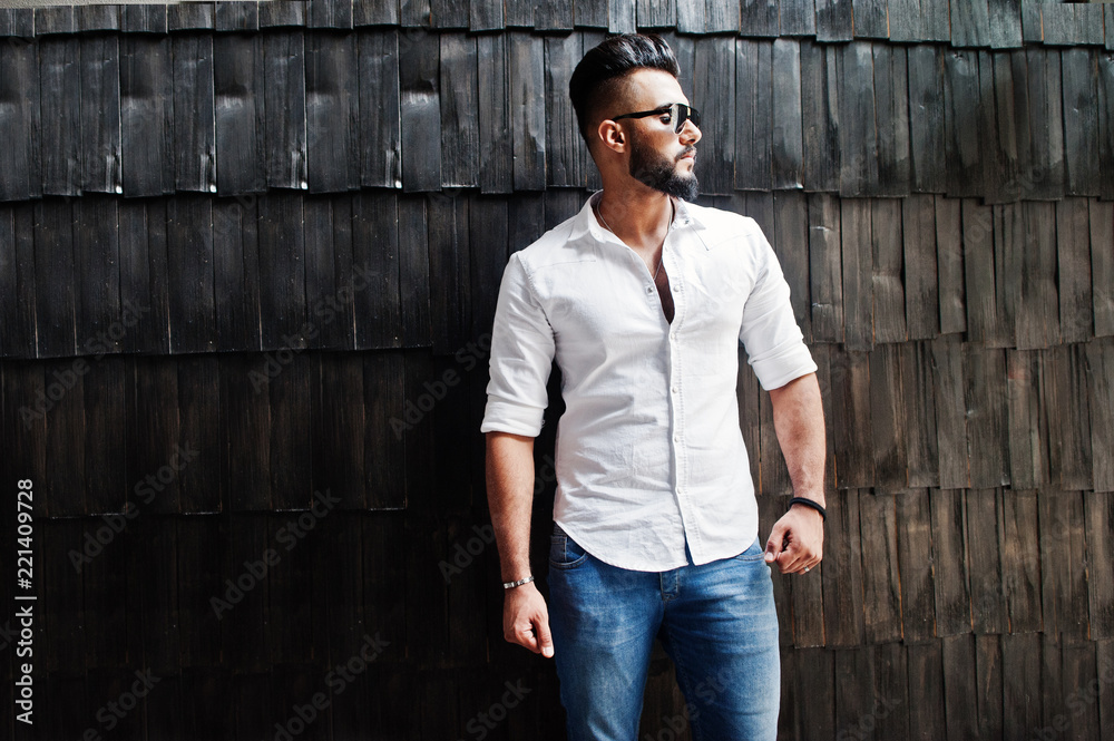 Obraz premium Stylish tall arabian man model in white shirt, jeans and sunglasses posed against wooden wall indoor. Beard attractive arab guy.