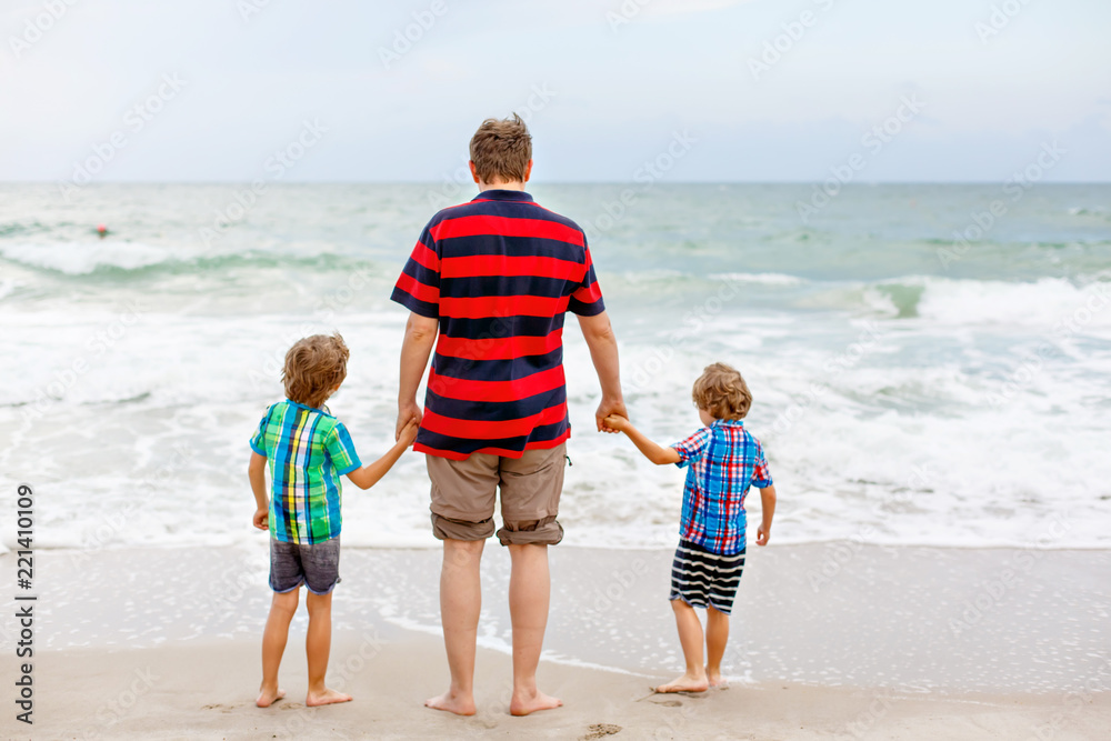 Two happy little kids boys and father standing on the beach of ocean and looking on horizon on stormy day. Family, dad and cute sons making vacations, dreaming and enjoying summer Two happy little kids boys and father standing on the beach of ocean and looking on horizon on stormy day. Family, dad and cute sons making vacations, dreaming and enjoying summer