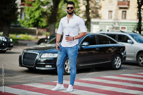 Stylish tall arabian man model in white shirt, jeans and sunglasses posed at street of city. Beard attractive arab guy with cup of coffee against black business car. © AS Photo Family