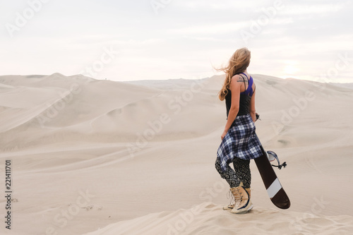 beautiful slender blond girl stands with snowboard against the background of the desert, dunes Sandboarding