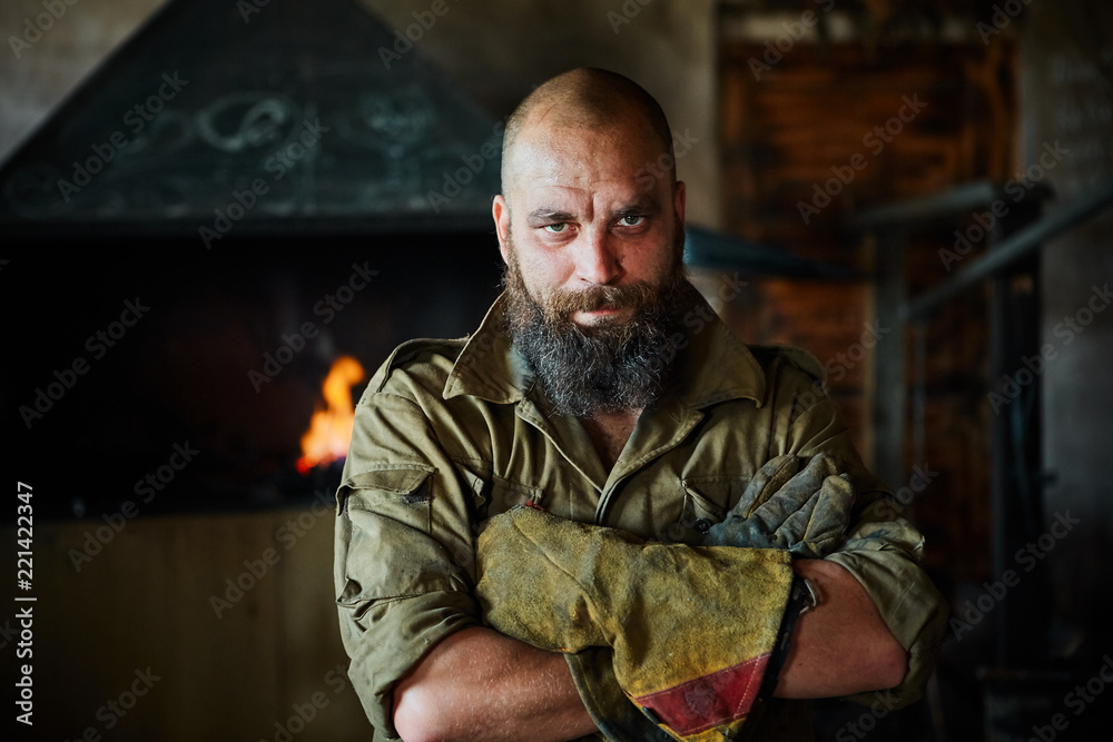 Portrait of a brutal, confident blacksmith, a man with a beard. Standing in the workshop, folding his arms over his chest