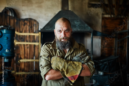 Portrait of a brutal, confident blacksmith, a man with a beard. Standing in the workshop, folding his arms over his chest