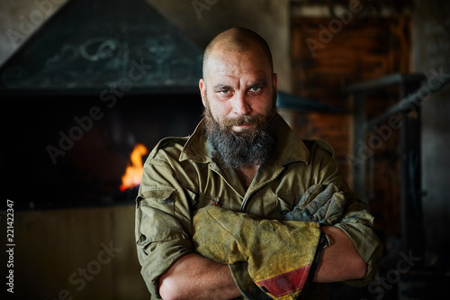 Portrait of a brutal, confident blacksmith, a man with a beard. Standing in the workshop, folding his arms over his chest