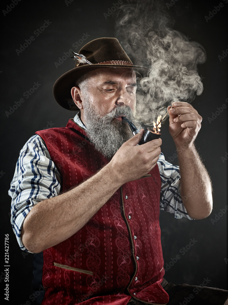 dramatic portrait of gray bearded senior man in hat smoking tobacco pipe.  view of Austrian, Tyrolean, Bavarian old man in national traditional  costume in retro style. Stock Photo | Adobe Stock