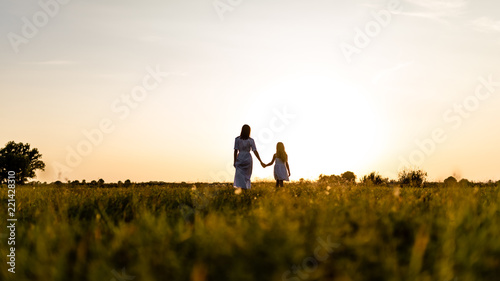 rear view of mother and daughter walking in green field with sunset on background