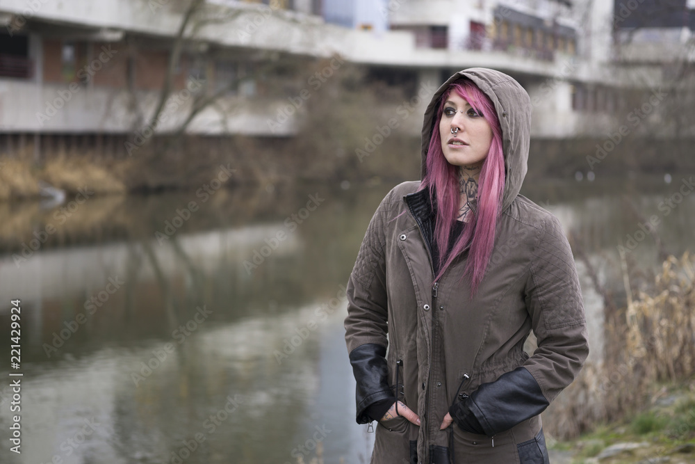 portrait of a young woman with pink hair standing by a river in bleak surroundings                          