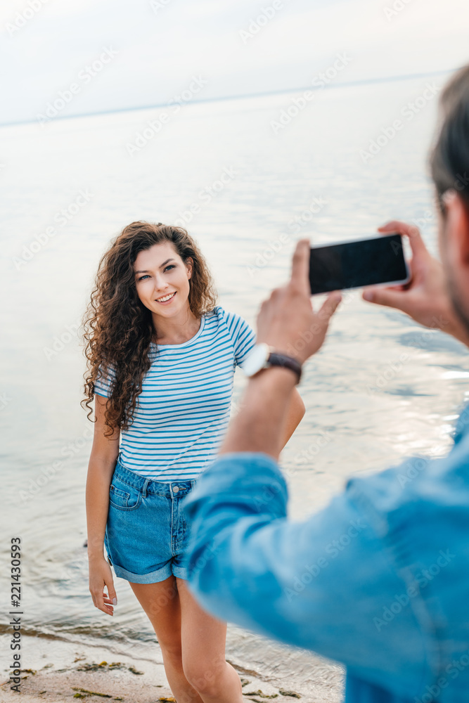 man taking photo of girlfriend near sea on smartphone