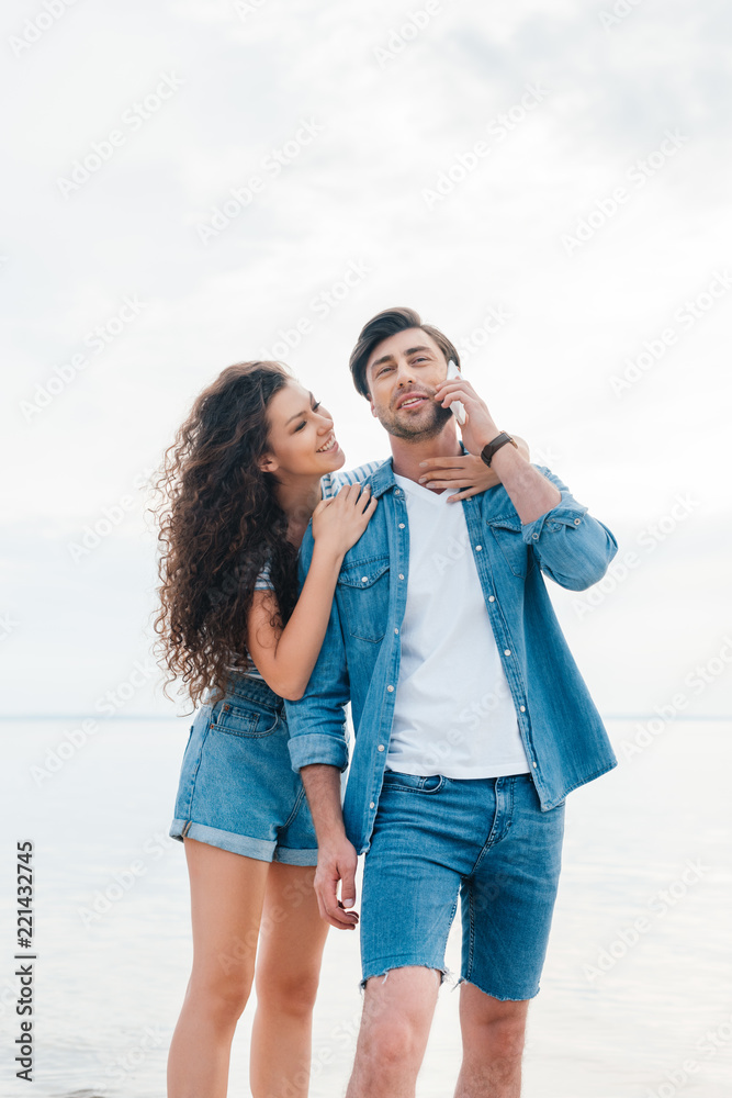 young girlfriend hugging her boyfriend while he talking on smartphone near sea