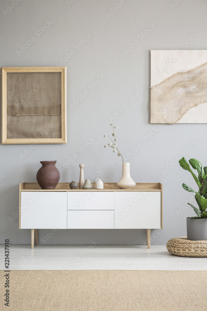 Fototapeta premium White wooden cupboard with decor and vase with flowers standing in real photo of bright room interior with grey wall and posters with fabrics