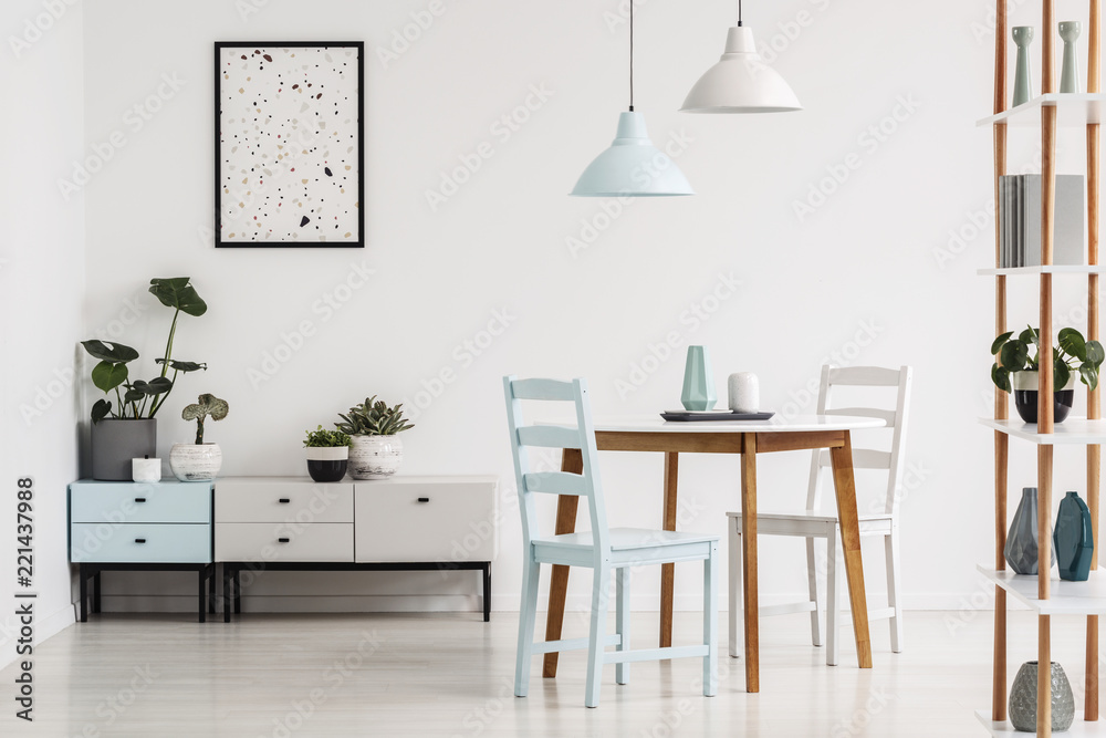 Lamps above wooden table and chairs in white dining room interior with poster above cupboard. Real photo Lamps above wooden table and chairs in white dining room interior with poster above cupboard. Real photo
