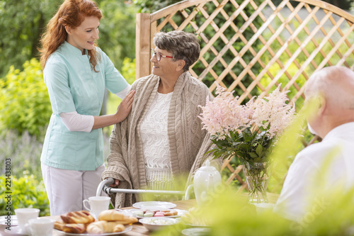 A tender caretaker assisting an elderly woman with a walker during an afternoon snack time on a patio in the garden of a private nursing home. photo