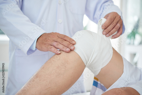 Unrecognizable medical practitioner wrapping bandage around knee of crop patient in doctor's office