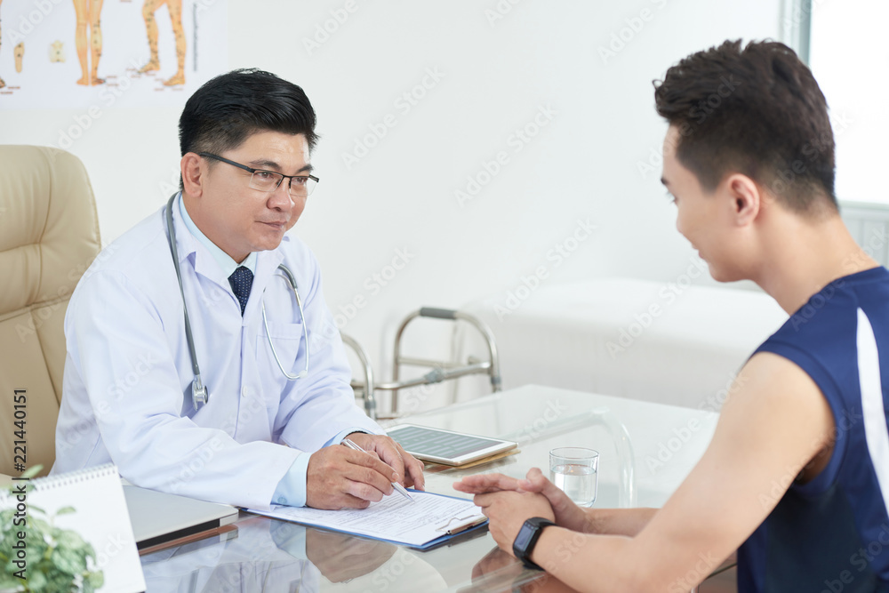 Side view of young guy sitting near table in doctor's office and consulting with adult Asian man in medical uniform Side view of young guy sitting near table in doctor's office and consulting with adult Asian man in medical uniform