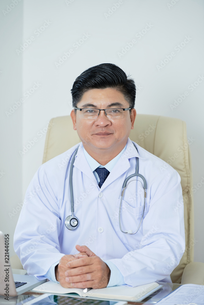 Confident Asian man in medical apparel looking at camera while sitting at table in doctor's office Confident Asian man in medical apparel looking at camera while sitting at table in doctor's office
