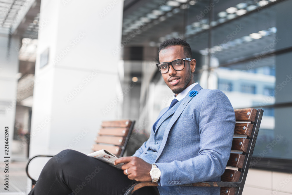 Obraz premium black man businessman in a business suit and glasses sits on a bench and reads a newspaper against the backdrop of a modern city