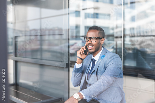 black man businessman in a business suit, expensive watch and glasses talking on the phone, decides business matters against the backdrop of a modern city to work