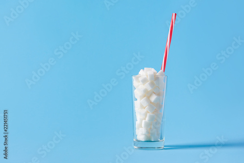 Glass full of sugar cubes with straw isolated on blue background. Unhealthy diet concept. Copy space