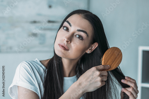 young brunette woman combing hair with hairbrush and looking at camera