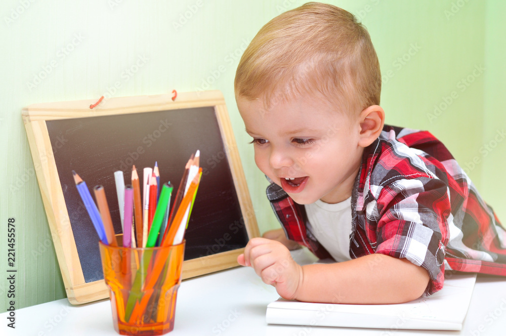 Smiling cute baby boy chooses color pencils and markers. A glass with colored pencils and markers. A two-year-old baby boy in a red checkered shirt sits at a table Smiling cute baby boy chooses color pencils and markers. A glass with colored pencils and markers. A two-year-old baby boy in a red checkered shirt sits at a table
