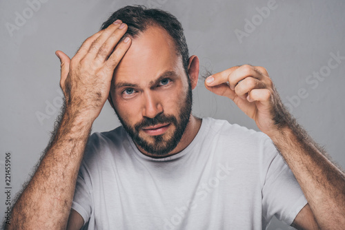 bearded middle aged man holding fallen hair and looking at camera isolated on grey photo