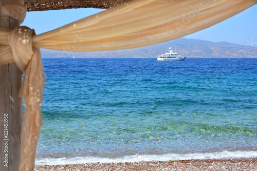 Fototapeta Naklejka Na Ścianę i Meble -  White ship in the sea visible through beach umbrella from the shore in Hydra island, Greece
