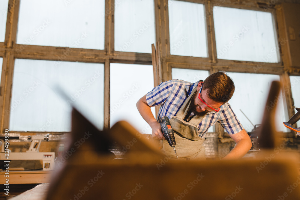 a bearded man in safety glasses and apron works as a drill in a carpenter's workshop a bearded man in safety glasses and apron works as a drill in a carpenter's workshop