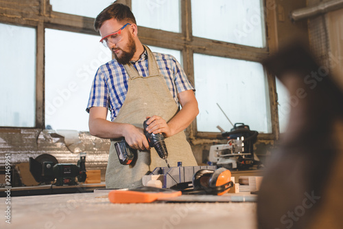 a bearded man in safety glasses and apron works as a drill in a carpenter's workshop