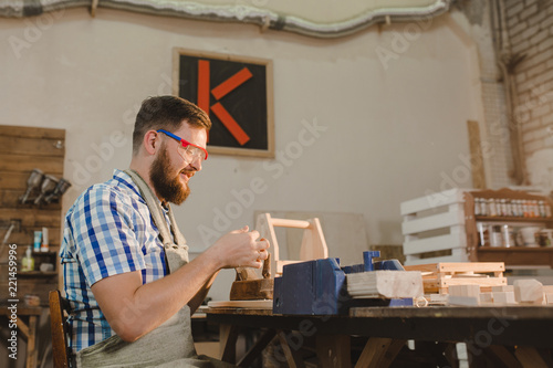 a bearded man in safety glasses and an apron works in a carpentry shop