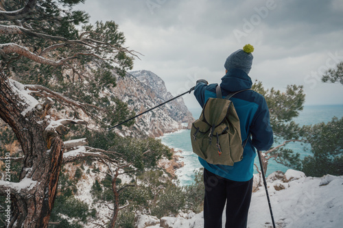Man with a backpack looks into the distance to the sea waves from the mountain. Hiking in the mountains in winter. Cold weather, snow. Winter trekking. Hiker in hike clothes. Back view. Backpacker.