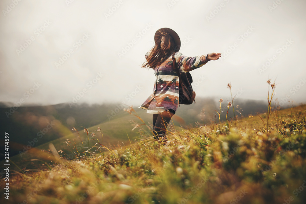 happy traveler hipster girl in hat, walking with backpack and smiling in windy mountains in clouds. space for text. amazing atmospheric emotional moment. travel and wanderlust concept - obrazy, fototapety, plakaty 