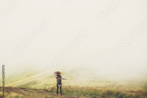 travel and wanderlust concept. stylish traveler girl holding hat, with backpack and windy hair, walking in mountains in clouds. summer vacation. space for text. amazing atmospheric moment