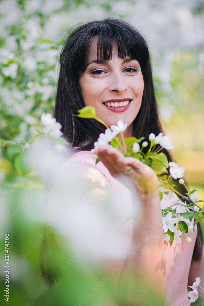 A beautiful girl with long black hair in a pink dress is walking in an apple orchard surrounded by flowers.. A model with clean skin. White flowers on the trees. A beautiful girl with long black hair in a pink dress is walking in an apple orchard surrounded by flowers.. A model with clean skin. White flowers on the trees.