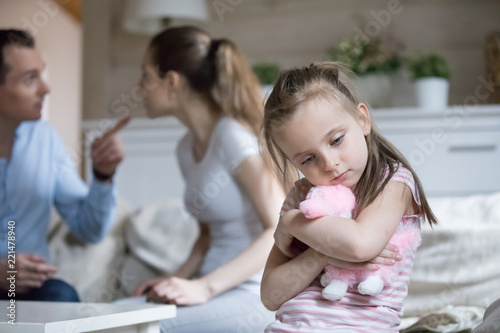 Sad little girl hug toy upset with parents fighting, frustrated small girl feel alone and depressed, mom and dad argue, lonely kid lack love and support. Psychology, family conflict, marriage break up