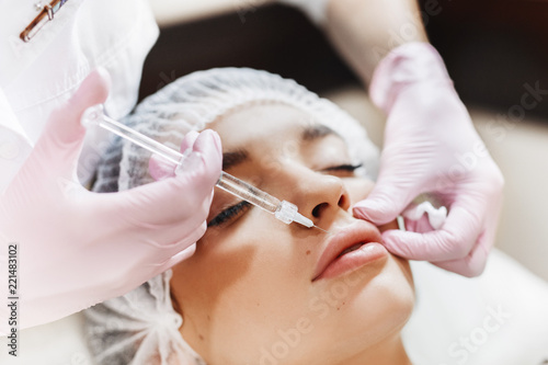 The process of lips enhancement. Cosmetician makes an injection of hyaluronic acid in a beautiful lips. The young girl with a beautiful face in the special hat and hands of the doctor in the pink glov