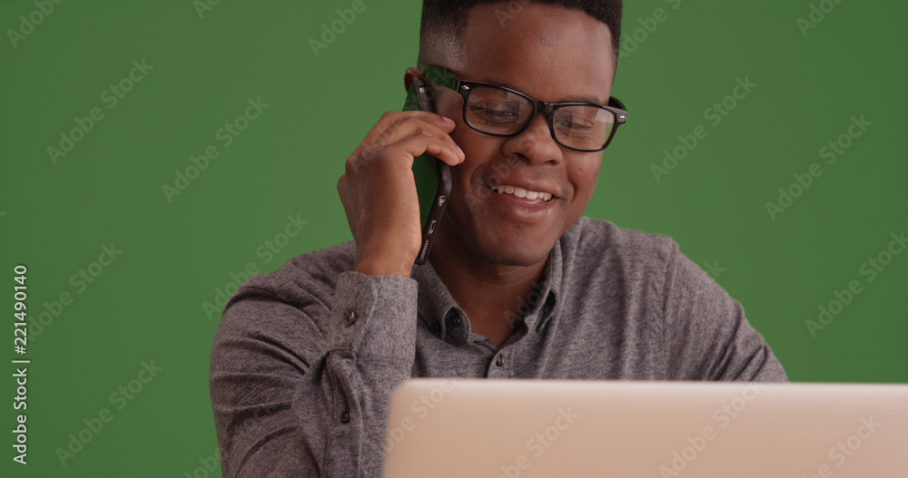 Black male with glasses talks on the phone working on his laptop on green screen Black male with glasses talks on the phone working on his laptop on green screen