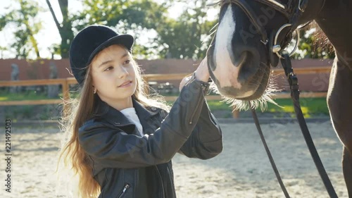 Cute girl in hat comes and caresses black mare's head on sunny background photo