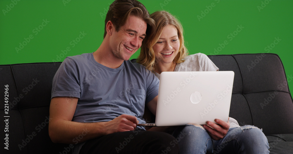 White millennial couple watching funny video on laptop on couch on green screen