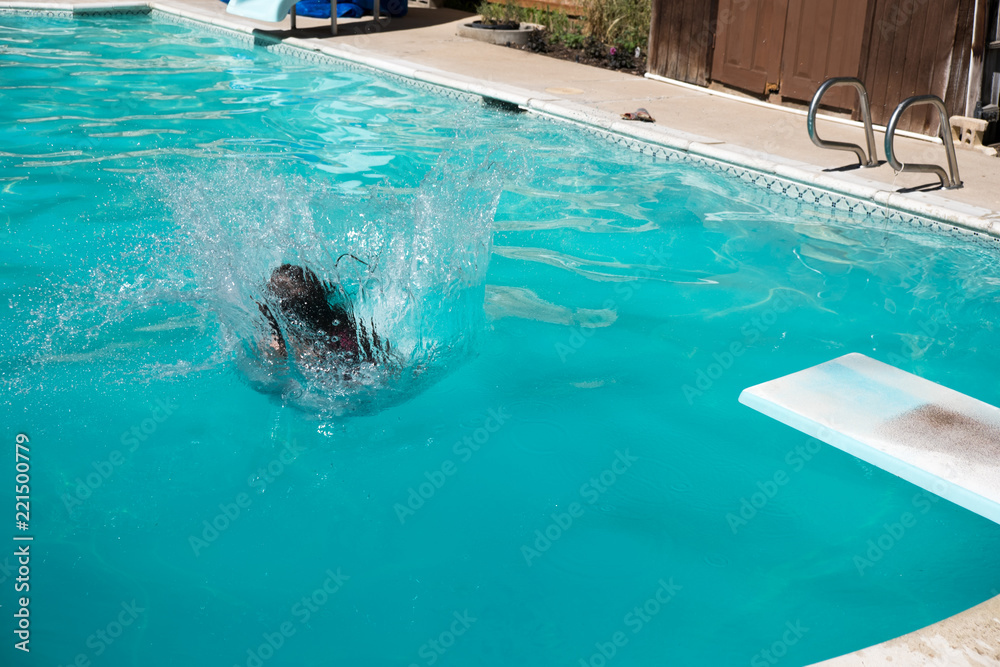 Dark haired woman doing a cannon ball into an outdoor swimming pool in a sunny summer day. Young woman jumps off diving board into a backyard swimming pool in the summer.